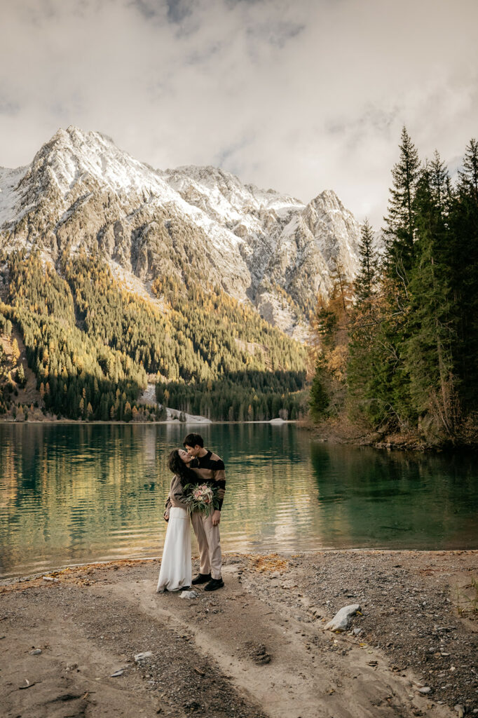 Couple kissing by mountain lake with snowy peaks.