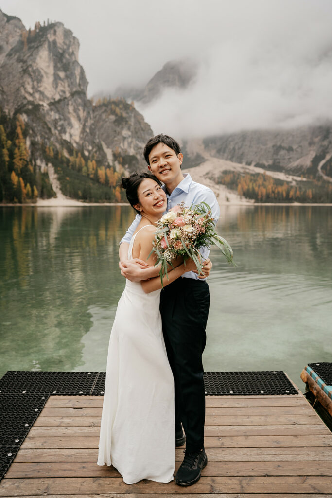 Couple embraces on lakeside dock, mountain view behind.