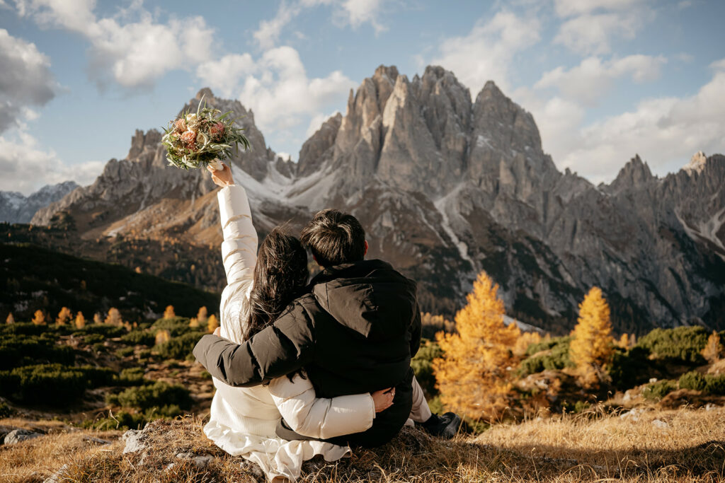 Couple embraces, holds flowers, with stunning mountain view.