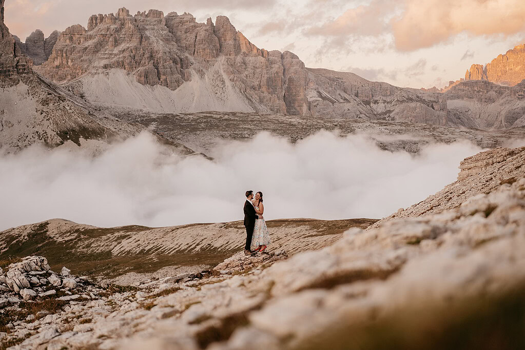 Couple embracing on mountain with clouds and peaks.