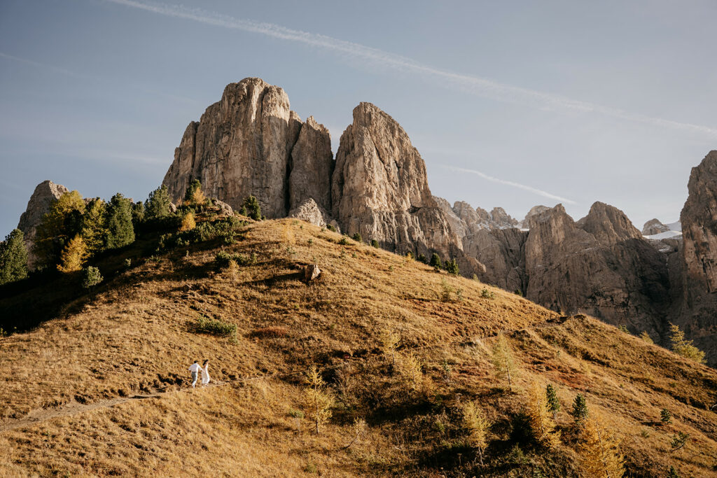 Couple hiking on mountain trail, rocky peaks behind.