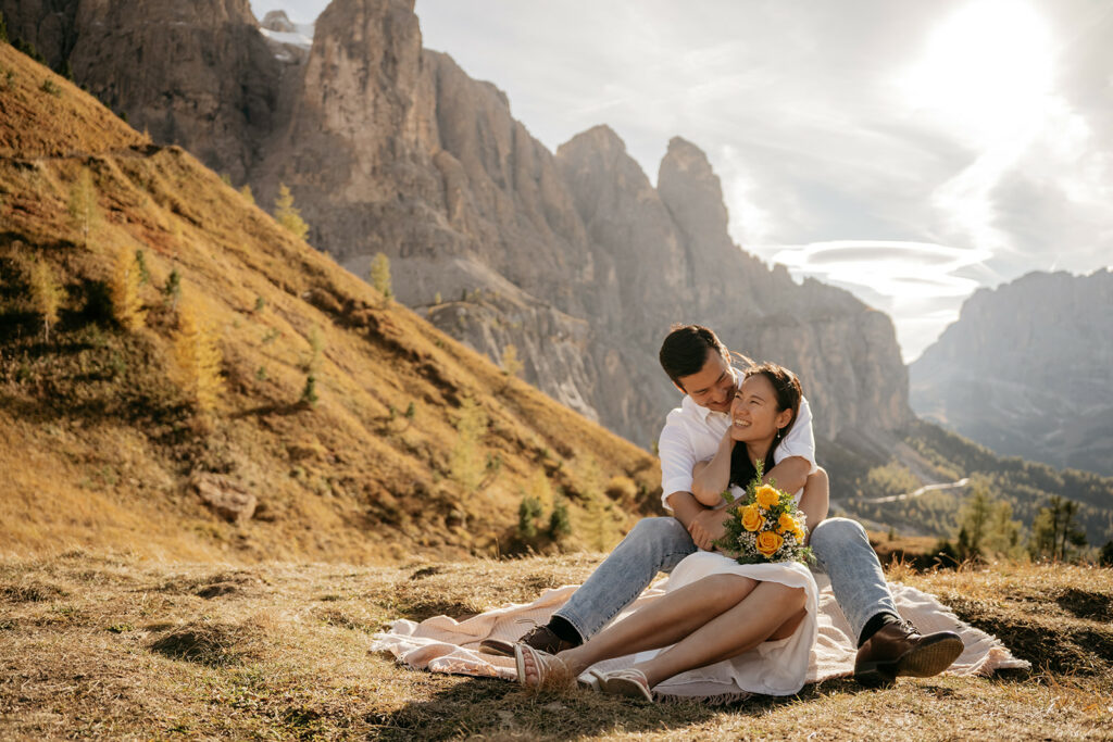 Couple embracing on mountain, smiling, holding flowers.