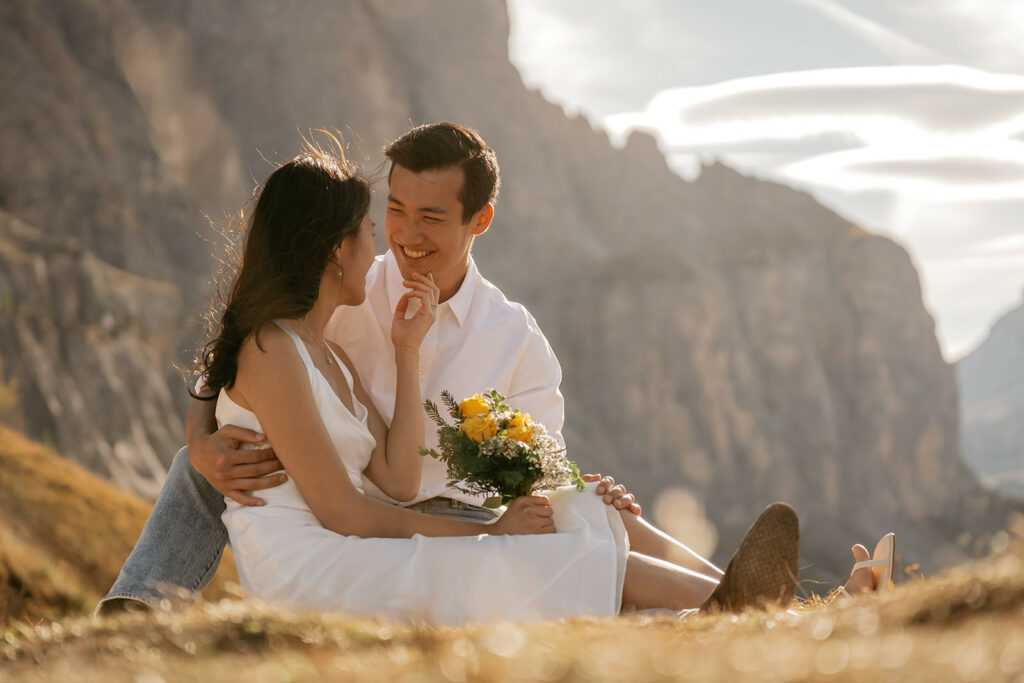 Couple sitting in mountains with flowers, smiling happily.