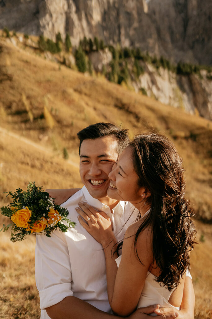 Couple smiling with yellow flowers in mountain background.