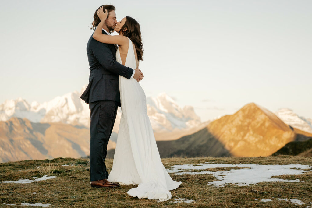 Bride and groom kiss on mountain top at sunset.