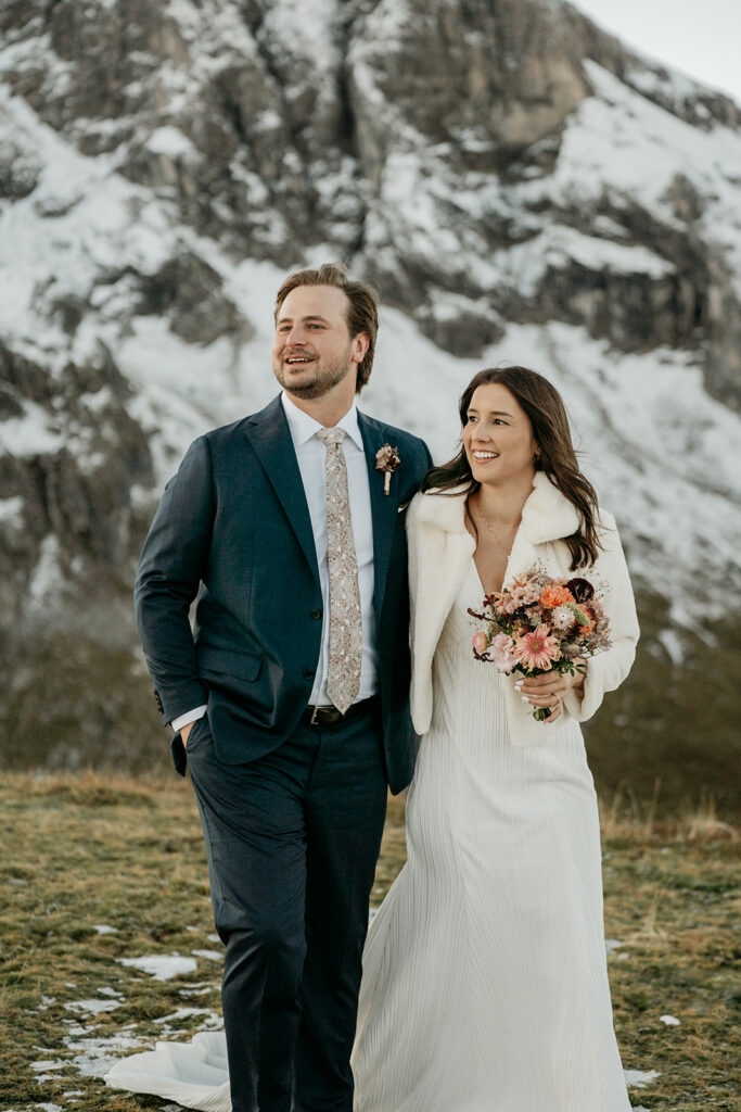 Couple in wedding attire standing before snowy mountain.