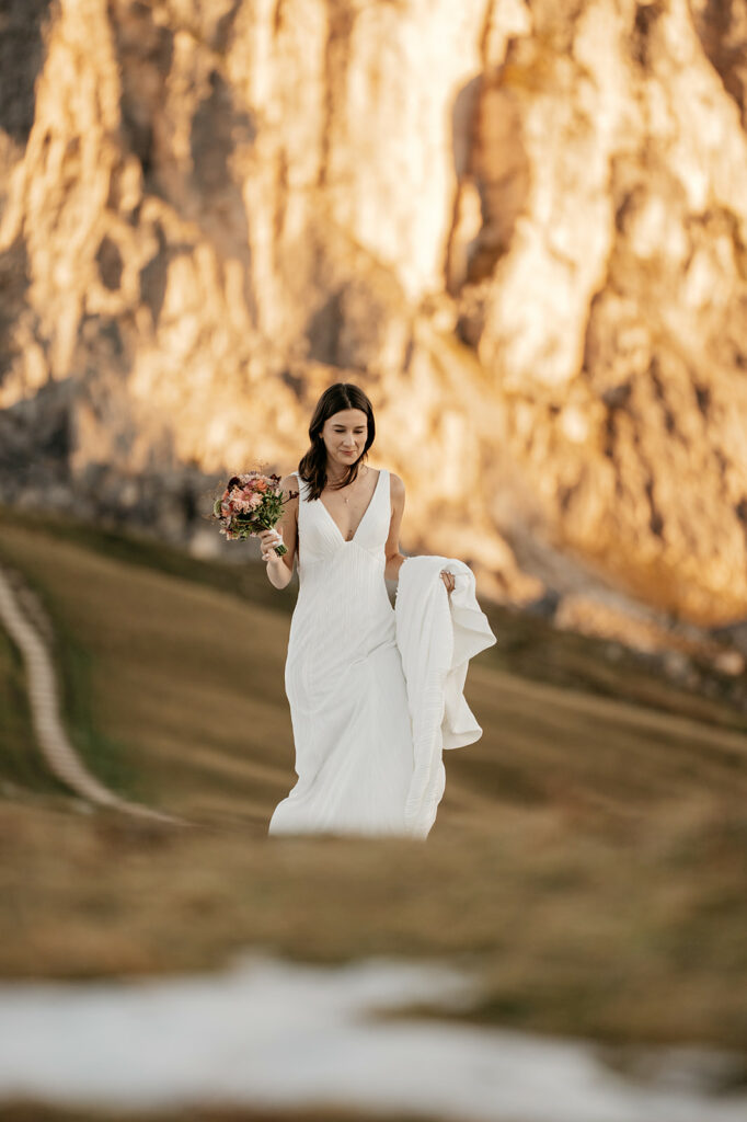 Bride walking on grassy mountain path with flowers.