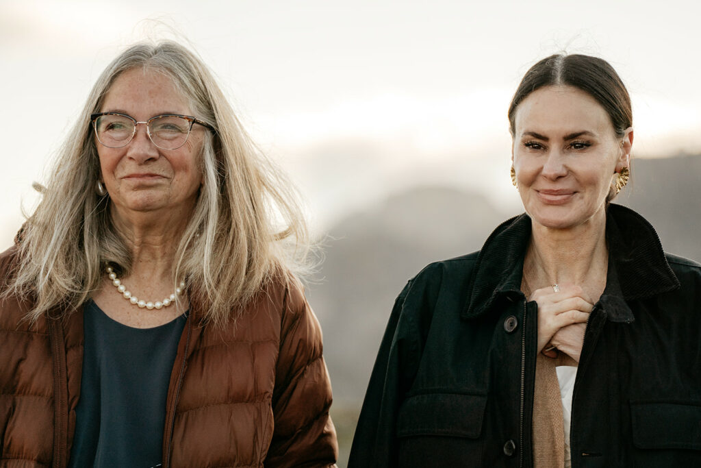 Two women smiling outdoors with mountains in background.