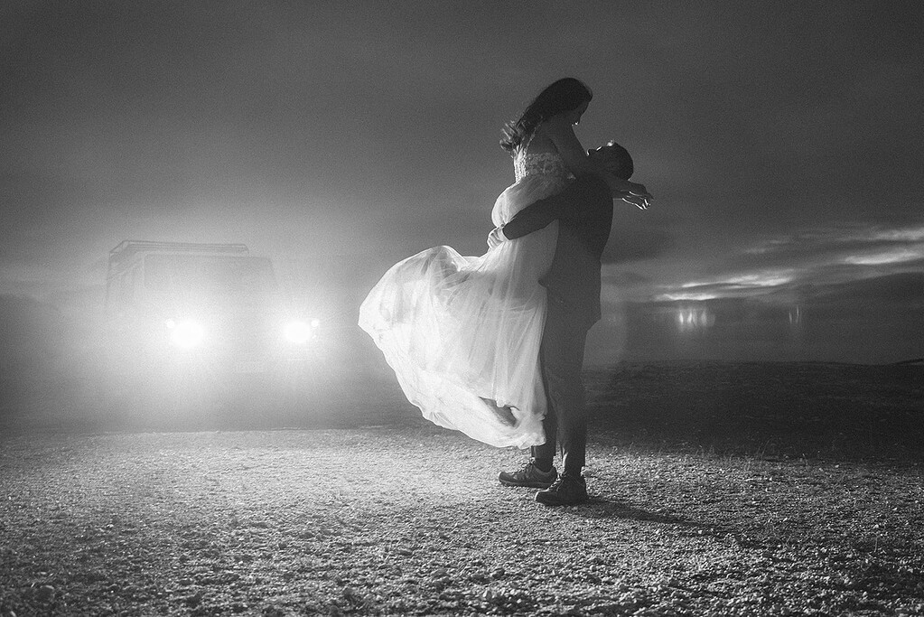 Couple embraced at night near vehicle with headlights.