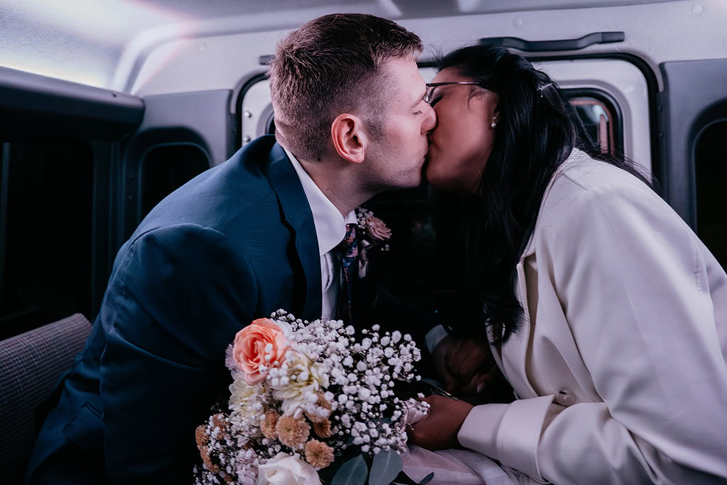Couple kissing in wedding attire, holding bouquet.