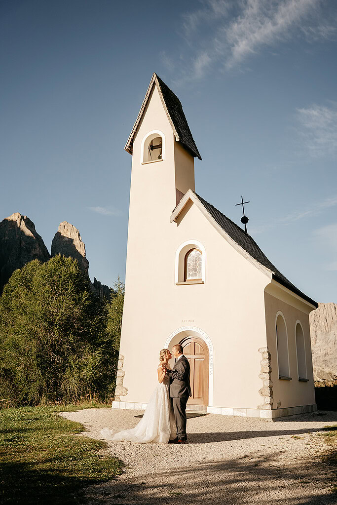 Couple embraces by mountain chapel under clear sky.
