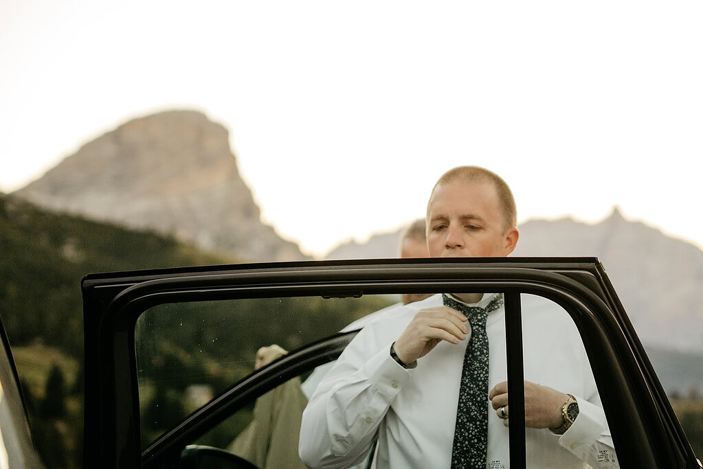 Man adjusts tie near car in mountain setting.